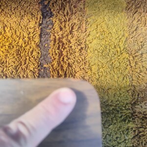 May include: A smooth, dark brown wooden surface with a rounded corner. The wood grain is visible across the flat surface, which is positioned against a textured, shaggy rug featuring vertical stripes of mustard yellow, brown, and dark green.