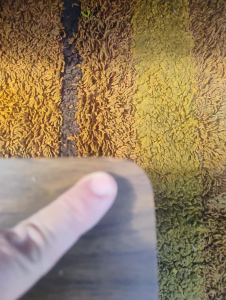 May include: A smooth, dark brown wooden surface with a rounded corner. The wood grain is visible across the flat surface, which is positioned against a textured, shaggy rug featuring vertical stripes of mustard yellow, brown, and dark green.