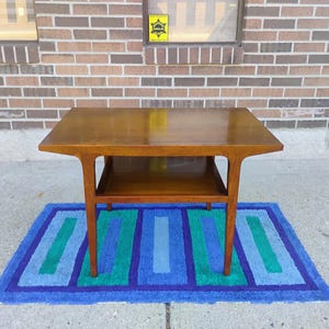 Puede incluir: Una mesa de centro de madera de estilo moderno de mediados de siglo con un estante inferior. La mesa tiene un acabado marrón oscuro y se encuentra sobre una alfombra de rayas azules y verdes.