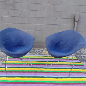 May include: Two blue, bowl-shaped chairs with rounded, shell-like seats and thin, silver-colored metal wire legs. The chairs feature a textured fabric upholstery and a curved, open design, positioned side-by-side on a flat, horizontal surface.