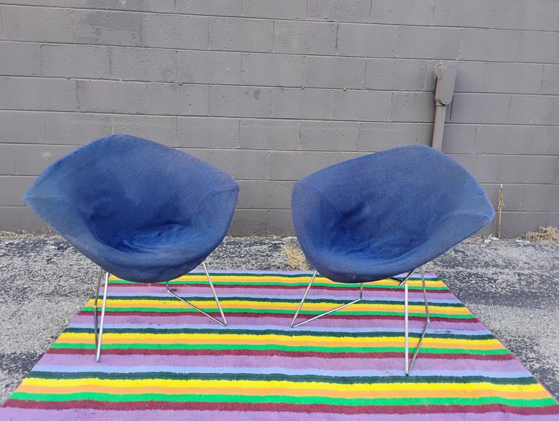May include: Two blue, bowl-shaped chairs with rounded, shell-like seats and thin, silver-colored metal wire legs. The chairs feature a textured fabric upholstery and a curved, open design, positioned side-by-side on a flat, horizontal surface.