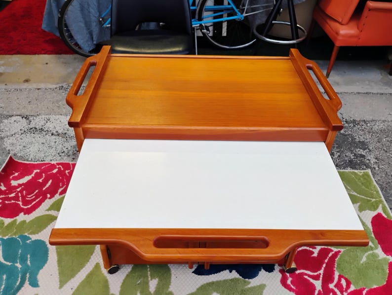 May include: A wooden serving cart with a white top and two handles. The cart has a lower shelf and wheels.