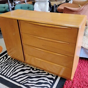 Op de afbeelding: Een licht houten dressoir met vier lades en een kastdeur. Het dressoir heeft een gebogen bovenkant en afgeronde randen. Het dressoir staat op een zwart-wit zebrakleurig tapijt.
