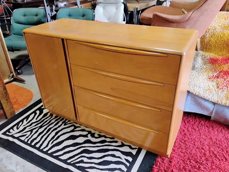 Op de afbeelding: Een licht houten dressoir met vier lades en een kastdeur. Het dressoir heeft een gebogen bovenkant en afgeronde randen. Het dressoir staat op een zwart-wit zebrakleurig tapijt.