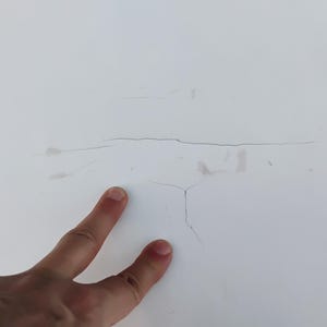 May include: A smooth white surface featuring a prominent horizontal crack and a smaller vertical branching crack. The white material shows subtle signs of wear with faint gray markings near the center of the damaged area.