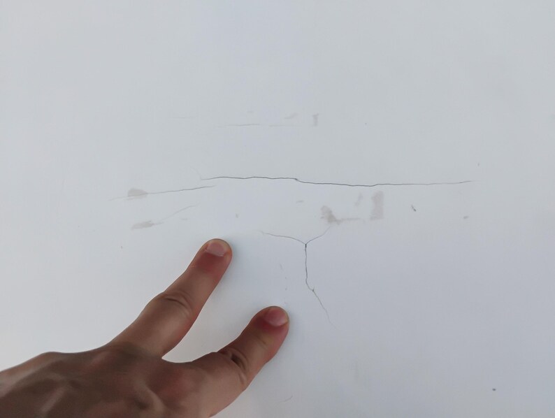 May include: A smooth white surface featuring a prominent horizontal crack and a smaller vertical branching crack. The white material shows subtle signs of wear with faint gray markings near the center of the damaged area.