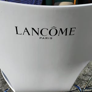 K&ouml;nnte beinhalten: Ein wei&szlig;er Stuhl mit geschwungener R&uuml;ckenlehne. Der schwarze Text "LANC&Ocirc;ME PARIS" ist auf der R&uuml;ckseite des Stuhls aufgedruckt. Der Stuhl hat ein silbernes Metallgestell.