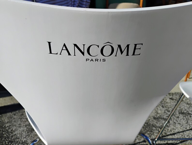 K&ouml;nnte beinhalten: Ein wei&szlig;er Stuhl mit geschwungener R&uuml;ckenlehne. Der schwarze Text "LANC&Ocirc;ME PARIS" ist auf der R&uuml;ckseite des Stuhls aufgedruckt. Der Stuhl hat ein silbernes Metallgestell.