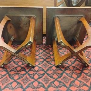 May include: Two wooden side tables shown upside down, each featuring a square top and a distinctive X-shaped base design. The underside of each table displays visible stamped text markings, and the legs are finished with small circular feet.