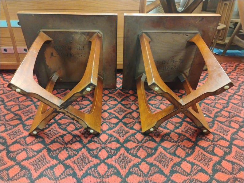 May include: Two wooden side tables shown upside down, each featuring a square top and a distinctive X-shaped base design. The underside of each table displays visible stamped text markings, and the legs are finished with small circular feet.