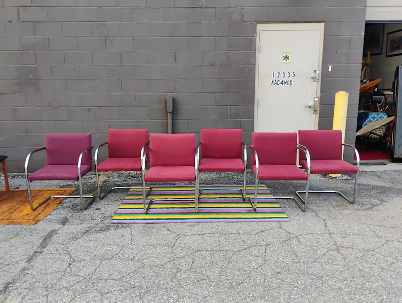 May include: A set of six cantilever chairs with polished chrome metal frames and upholstered seats and backrests in varying shades of red and magenta. Each chair features a continuous tubular metal base and integrated armrests with black caps.