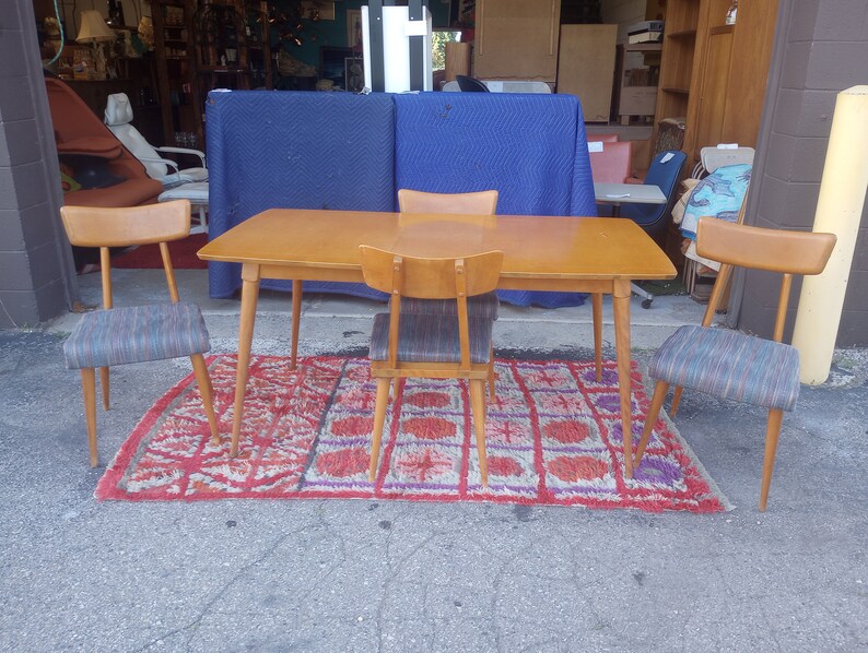 May include: A set of four mid-century modern dining chairs with a wood frame and patterned fabric seats surrounding a rectangular wood table with tapered legs. The chairs and table are on a red and purple patterned rug.