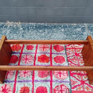May include: A rectangular, wooden bookshelf with a warm, reddish-brown finish. The shelf has two horizontal supports and two vertical side supports. It sits on a colorful, patterned rug with red, pink, and purple floral designs.