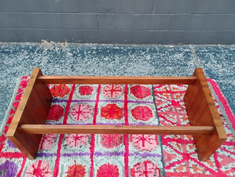 May include: A rectangular, wooden bookshelf with a warm, reddish-brown finish. The shelf has two horizontal supports and two vertical side supports. It sits on a colorful, patterned rug with red, pink, and purple floral designs.