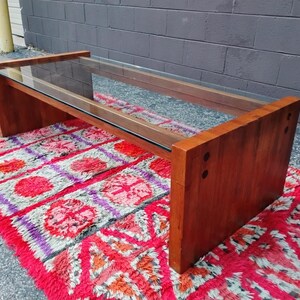 May include: A rectangular coffee table with a clear glass top and a wooden frame. The frame is made of dark brown wood. The table is placed on a red and pink patterned rug.