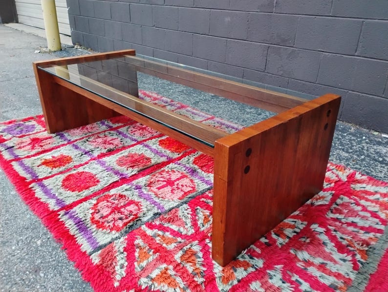 May include: A rectangular coffee table with a clear glass top and a wooden frame. The frame is made of dark brown wood. The table is placed on a red and pink patterned rug.