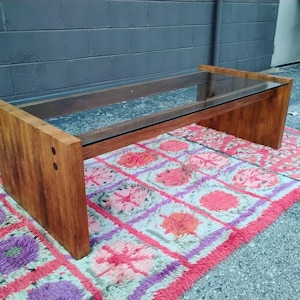 Puede incluir: Una mesa de centro rectangular con una tapa de cristal transparente y una base de madera. La base es de madera maciza de color marrón claro. La mesa está colocada sobre una alfombra colorida con diseños rosas, morados y naranjas.