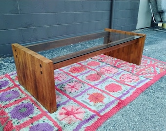 Könnte beinhalten: Ein rechteckiger Couchtisch mit einer transparenten Glasplatte und einem Holzsockel. Der Sockel besteht aus massivem, hellbraunem Holz. Der Tisch steht auf einem bunten Teppich mit rosa, lila und orangefarbenen Mustern.