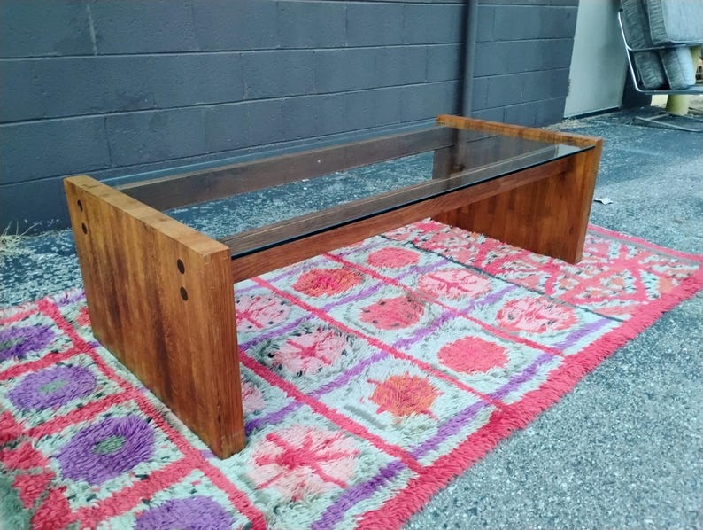 May include: A rectangular coffee table with a clear glass top and a wooden base. The base is made of solid, light brown wood. The table is placed on a colorful rug with pink, purple, and orange patterns.