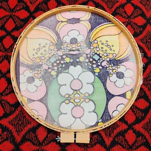 May include: A round wooden embroidery hoop holding a fabric with a floral design. The design features large flowers in pink, white, and green, with smaller colorful dots and yellow and orange petal shapes. The background is dark blue.