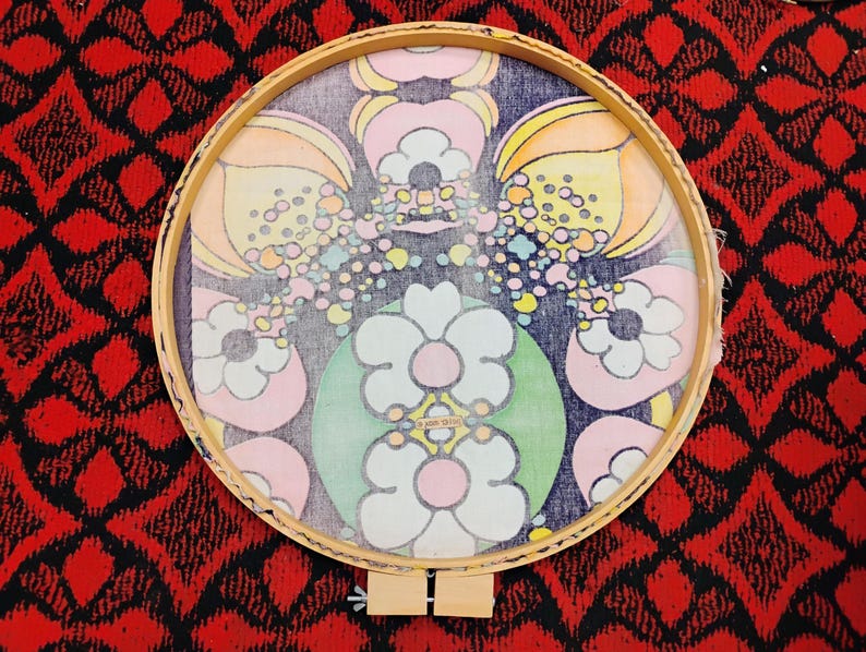 May include: A round wooden embroidery hoop holding a fabric with a floral design. The design features large flowers in pink, white, and green, with smaller colorful dots and yellow and orange petal shapes. The background is dark blue.