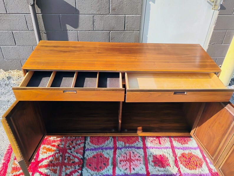 Op de afbeelding: Een rechthoekig houten dressoir met een warme bruine afwerking, voorzien van twee bovenste lades en twee onderste kastdeuren. De linkerlade bevat verdelers met een donkere stoffen voering, terwijl de rechterlade open en leeg is.