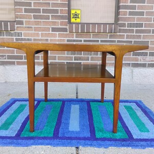 May include: A mid-century modern end table with a wood top and a lower shelf. The table has a unique, curved design and is made of solid wood. The table is sitting on a blue and green striped rug.