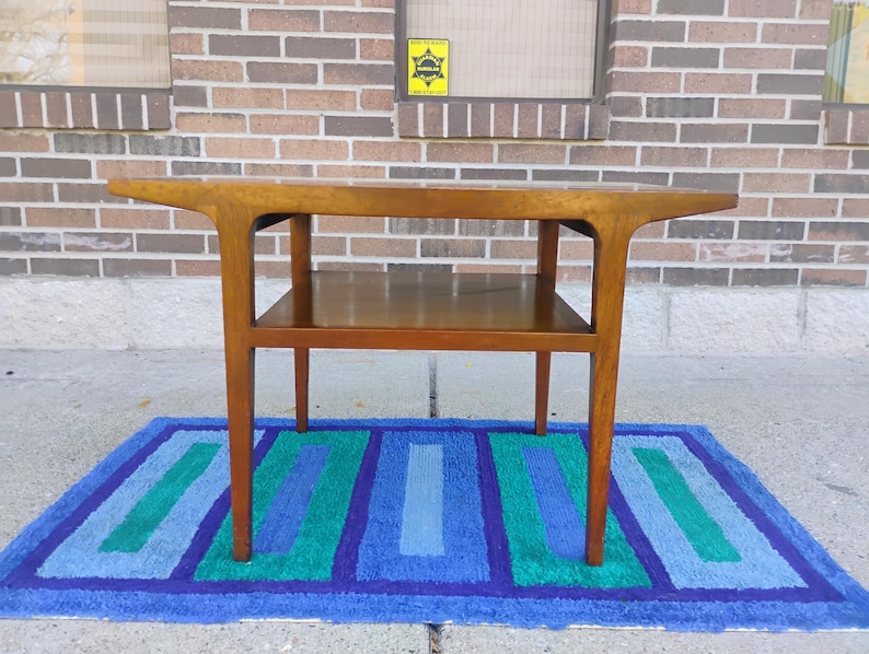 May include: A mid-century modern end table with a wood top and a lower shelf. The table has a unique, curved design and is made of solid wood. The table is sitting on a blue and green striped rug.