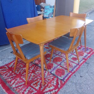 May include: A set of four mid-century modern dining chairs with a wood frame and a light wood table top. The chairs have a woven fabric seat and back. The table and chairs are on a red and purple rug.