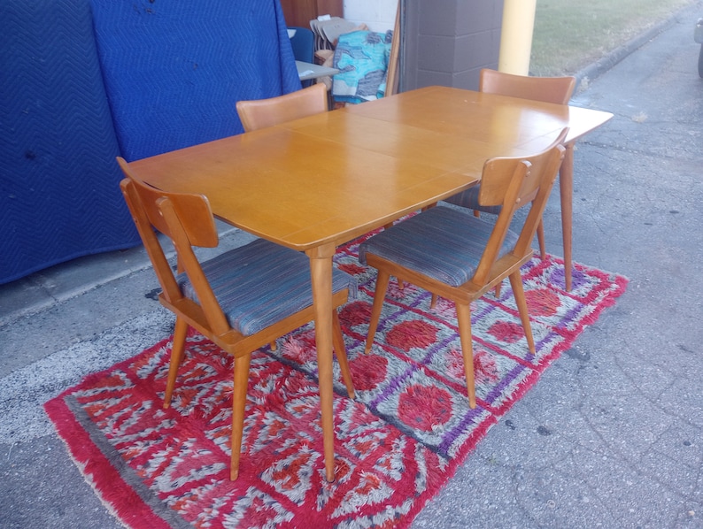 May include: A set of four mid-century modern dining chairs with a wood frame and a light wood table top. The chairs have a woven fabric seat and back. The table and chairs are on a red and purple rug.