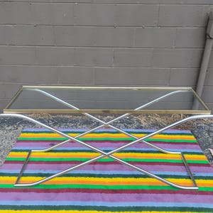 May include: A rectangular coffee table featuring a clear glass top with a thin brass-toned metal frame. The base is constructed from two intersecting silver-toned metal tubes that form an X-shape, providing structural support for the tabletop.