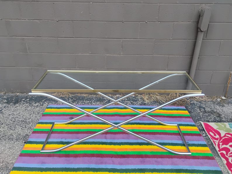 May include: A rectangular coffee table featuring a clear glass top with a thin brass-toned metal frame. The base is constructed from two intersecting silver-toned metal tubes that form an X-shape, providing structural support for the tabletop.
