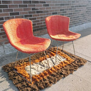May include: Two mid-century modern chairs with orange fabric seats and chrome legs. The chairs sit on a brown, orange, and white patterned rug. The chairs are positioned on a concrete surface next to a brick wall.