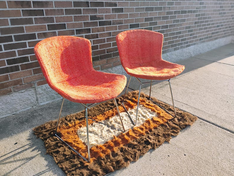 May include: Two mid-century modern chairs with orange fabric seats and chrome legs. The chairs sit on a brown, orange, and white patterned rug. The chairs are positioned on a concrete surface next to a brick wall.
