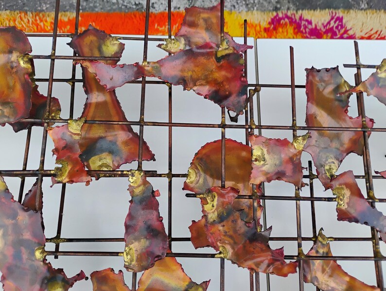 May include: Abstract wall art featuring a grid of metal with irregular, fragmented pieces of copper-toned metal with red and gold accents.