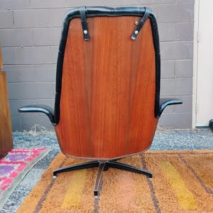 May include: A high-back swivel chair featuring a polished wood veneer back panel, black leather upholstery, and black leather straps secured with silver-toned metal hardware. The chair is supported by a four-pronged black metal base.