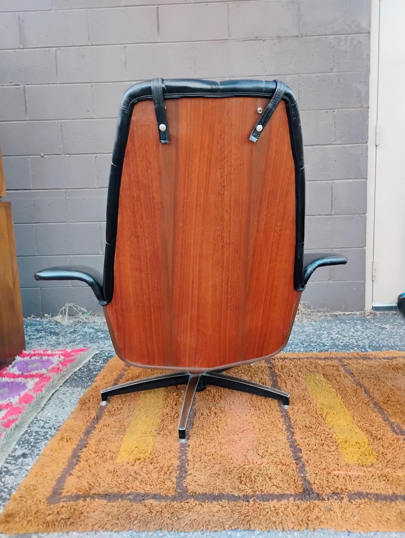 May include: A high-back swivel chair featuring a polished wood veneer back panel, black leather upholstery, and black leather straps secured with silver-toned metal hardware. The chair is supported by a four-pronged black metal base.