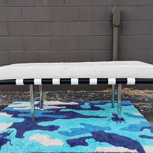 May include: A modern bench with a white cushioned seat and a black frame accented with white stripes. The bench has four chrome legs and sits on a blue, white, and navy camouflage rug.