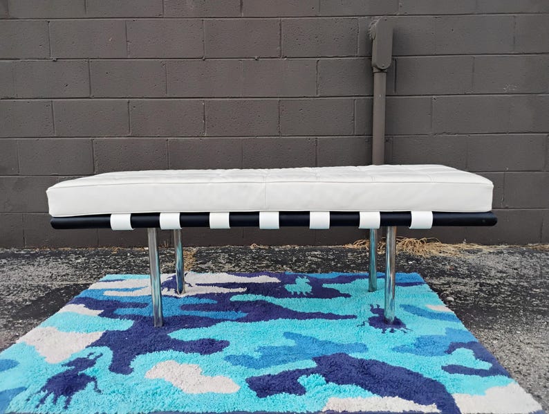 May include: A modern bench with a white cushioned seat and a black frame accented with white stripes. The bench has four chrome legs and sits on a blue, white, and navy camouflage rug.