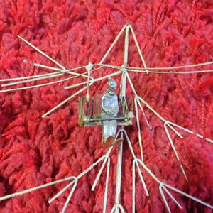 Pode incluir: Uma escultura em metal prateado de uma pessoa sentada, segurando uma bandeja, dentro de uma estrutura de arame. A escultura est&aacute; sobre um fundo vermelho vibrante e texturizado.