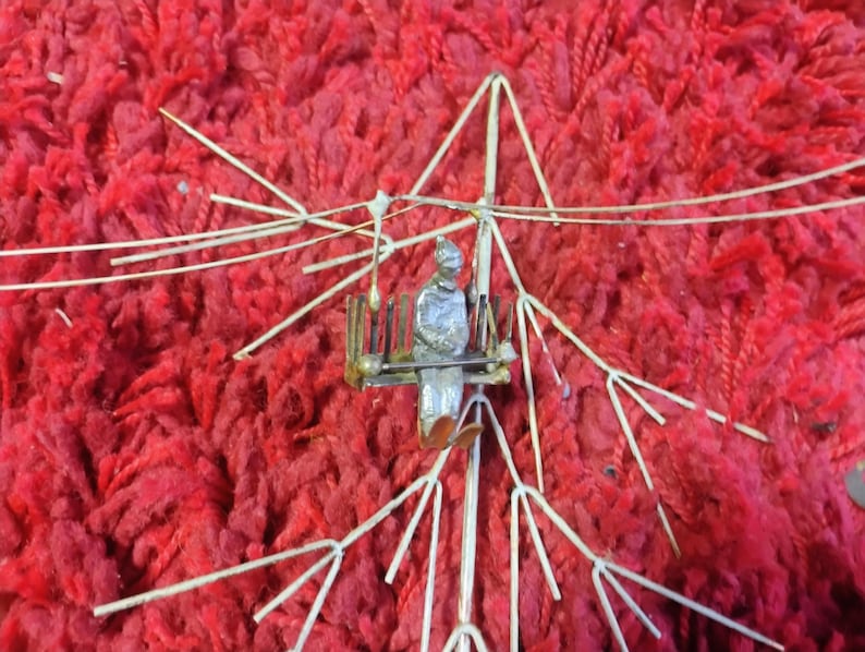 Pode incluir: Uma escultura em metal prateado de uma pessoa sentada, segurando uma bandeja, dentro de uma estrutura de arame. A escultura est&aacute; sobre um fundo vermelho vibrante e texturizado.