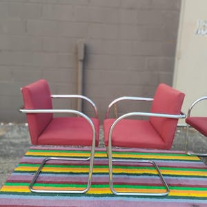 May include: Two armchairs featuring red upholstered seats and backrests supported by continuous tubular chrome frames. The chairs have a cantilevered design with curved armrests and a rectangular base structure, providing a stable seating arrangement.