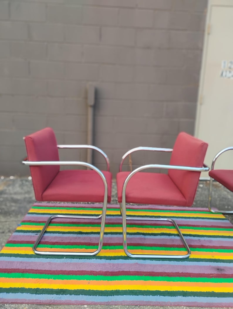 May include: Two armchairs featuring red upholstered seats and backrests supported by continuous tubular chrome frames. The chairs have a cantilevered design with curved armrests and a rectangular base structure, providing a stable seating arrangement.