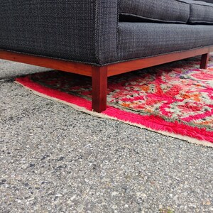 K&ouml;nnte beinhalten: Ein Sofa mit einem Bezug aus dunkelgrauem Stoff mit feinem wei&szlig;em Gittermuster, das &uuml;ber einen h&ouml;lzernen Unterrahmen mit geraden Beinen verf&uuml;gt. Das Sofa steht auf einem lebhaft strukturierten Teppich mit einem markanten geometrischen Muster in Pink, Rot und Orange.