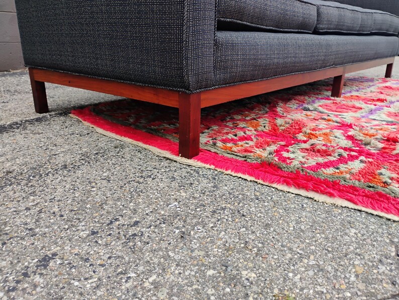 K&ouml;nnte beinhalten: Ein Sofa mit einem Bezug aus dunkelgrauem Stoff mit feinem wei&szlig;em Gittermuster, das &uuml;ber einen h&ouml;lzernen Unterrahmen mit geraden Beinen verf&uuml;gt. Das Sofa steht auf einem lebhaft strukturierten Teppich mit einem markanten geometrischen Muster in Pink, Rot und Orange.
