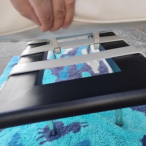 May include: A black and silver metal chair with a white seat. The chair has a rectangular seat and four silver legs. The chair is on a blue and white patterned rug. A hand is holding the seat.