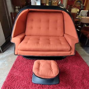 May include: A black and orange retro-style modular sofa with a matching ottoman. The sofa has a curved, enclosed back and a swivel base. The ottoman has a button-tufted top.
