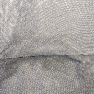 May include: A close-up view of a beige, woven fabric with a visible, textured grid pattern. The material features a neutral, light tan color with subtle variations in tone across the surface, highlighting the interlaced threads of the textile.