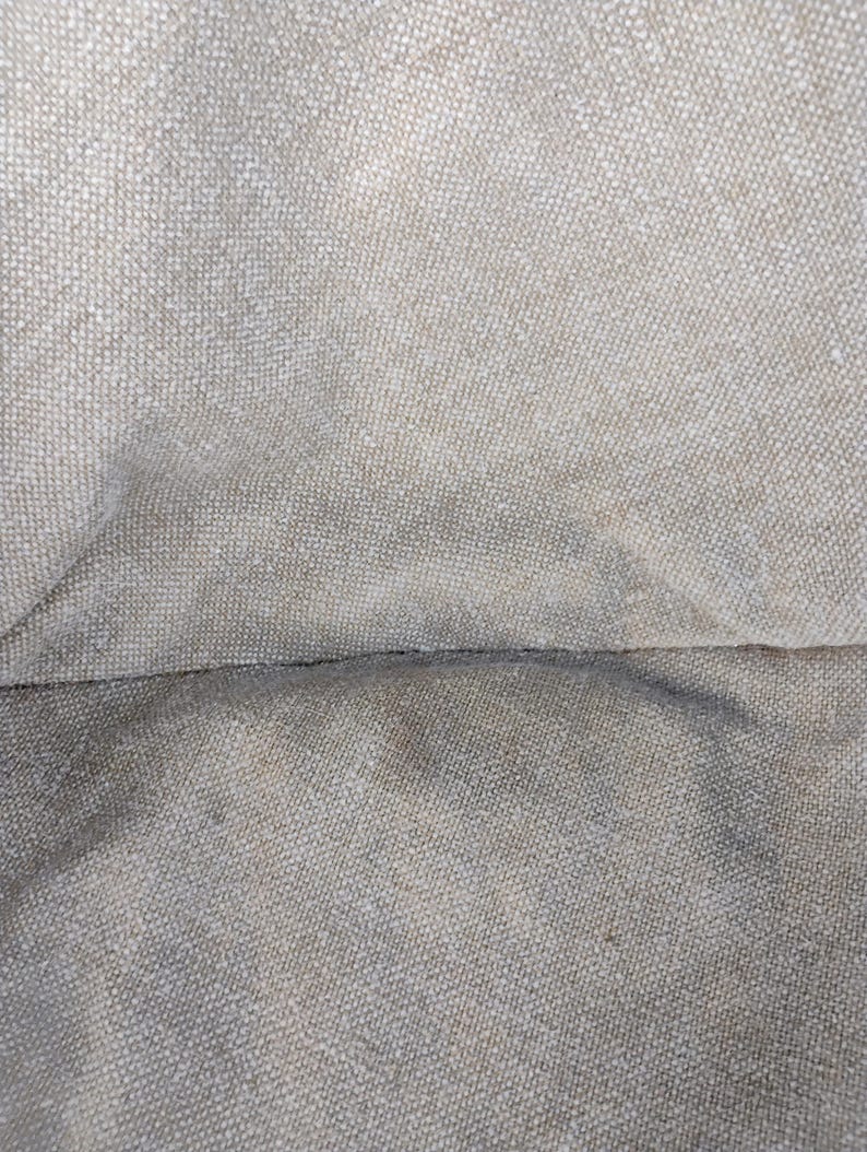 May include: A close-up view of a beige, woven fabric with a visible, textured grid pattern. The material features a neutral, light tan color with subtle variations in tone across the surface, highlighting the interlaced threads of the textile.