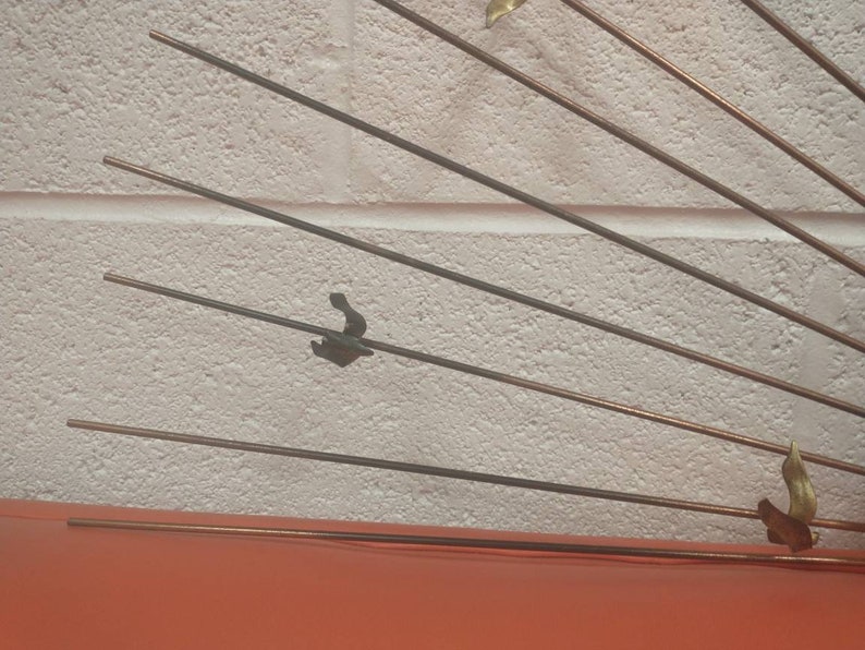 May include: A wall sculpture made of metal with a sunburst design. The sculpture has multiple thin metal rods radiating out from a central point. The rods are a mix of copper and black metal. There are small metal shapes attached to the ends of some of the rods.